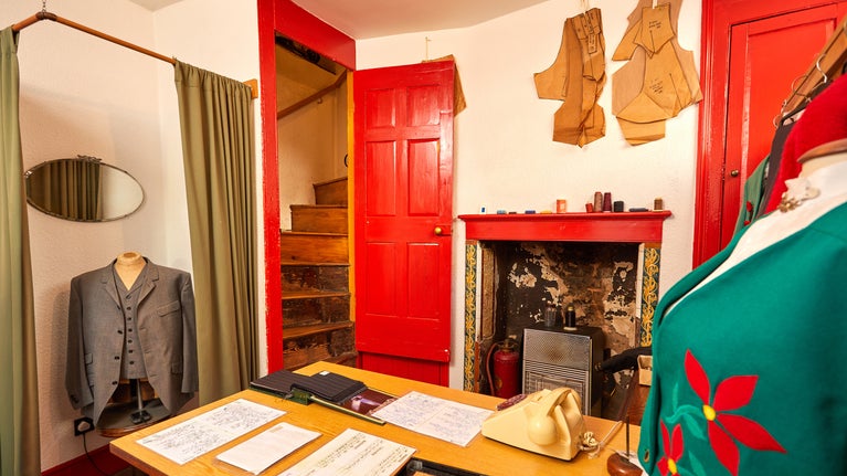 Inside George Saunders' tailor shop. A small space, tailor's dummies can be seen in the foreground and background, a table is between the tailor's dummies with an old dial telephone and a selection of documents.